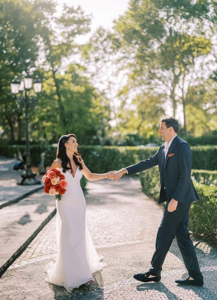 A bride in a white dress holds a bouquet of orange flowers, smiling and reaching out to a man in a dark suit. They are outdoors on a sunny day at Villa Cora, with trees and a path in the background, perfectly captured by the destination wedding photographer.