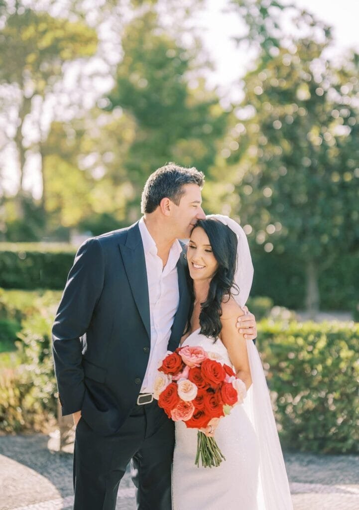 At a picturesque Villa Cora in Florence, a groom in a black suit kisses the forehead of his smiling bride, who holds a bouquet of red and pink roses. They stand outdoors amidst lush greenery on this sunny destination wedding day, perfectly captured by the talented photographer.