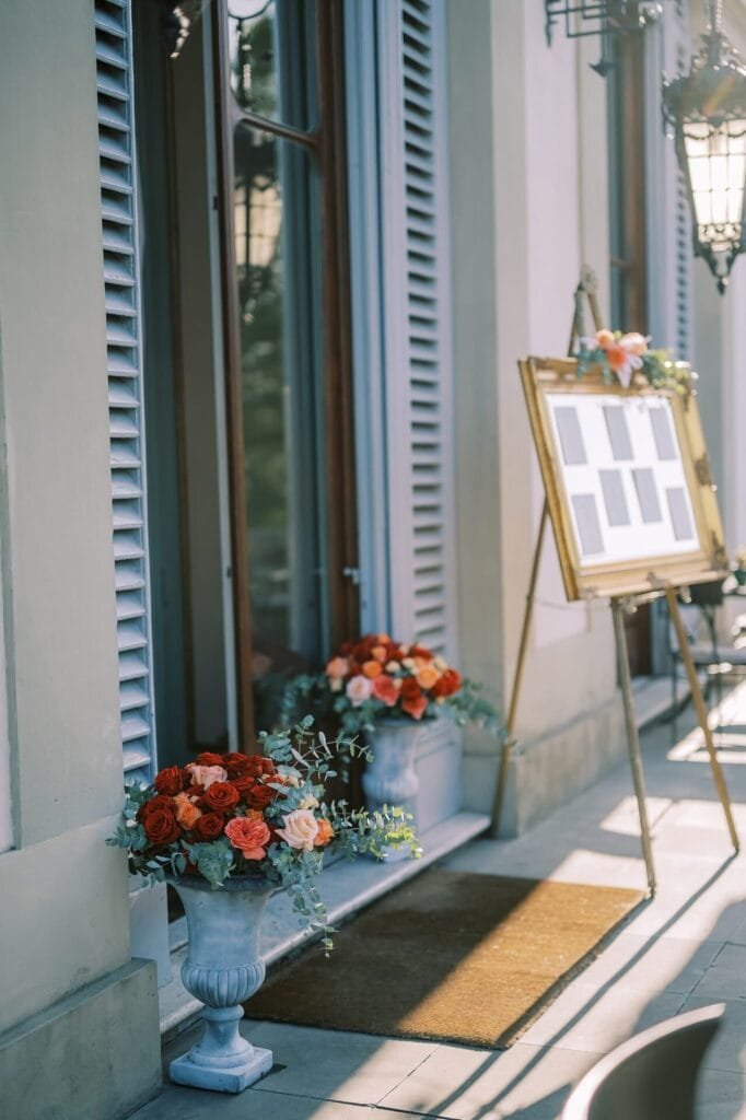 In the sunny elegance of Villa Cora in Florence, a framed board on an easel lists guest names for an exquisite destination wedding. Vibrant floral arrangements with red, orange, and pink roses sit beside shuttered windows, casting soft shadows perfect for a photographers lens.