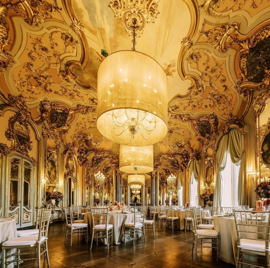 The ornate ballroom at Villa Cora, Florence, features elaborate golden details, large chandeliers, and decorated mirrors. Tables with white tablecloths and elegant chairs await an event. The intricate ceiling boasts sculpted designs while natural light filters in through tall windows—a perfect destination for a wedding photographer.