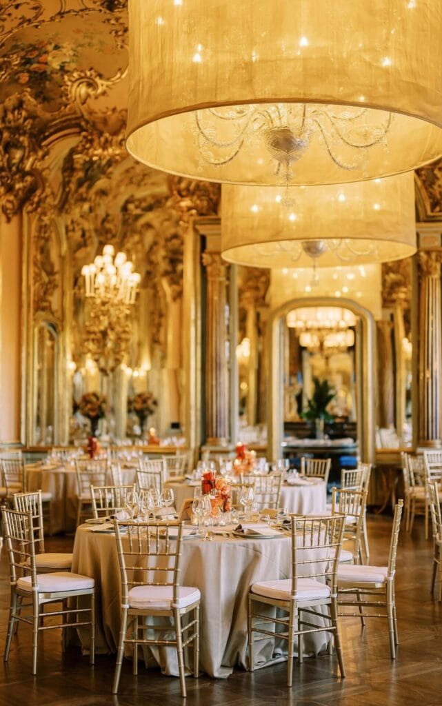 A luxurious ballroom at Villa Cora in Florence exudes elegance with ornate decor, large chandeliers, and mirrored walls. Elegant round tables featuring white tablecloths and fine dinnerware are surrounded by gold Chiavari chairs as soft lighting creates an inviting atmosphere for your destination wedding.