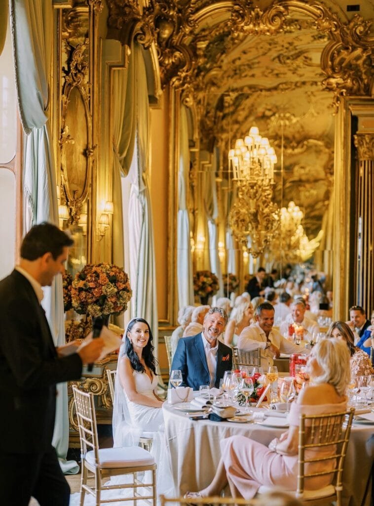 At a Villa Cora Florence destination wedding, the reception unfolds in an opulent, gold-adorned ballroom with elaborate chandeliers. Guests are seated at round tables with white tablecloths. The bride and groom smile as a man gives a speech. Large mirrors and floral arrangements enhance the elegant atmosphere.