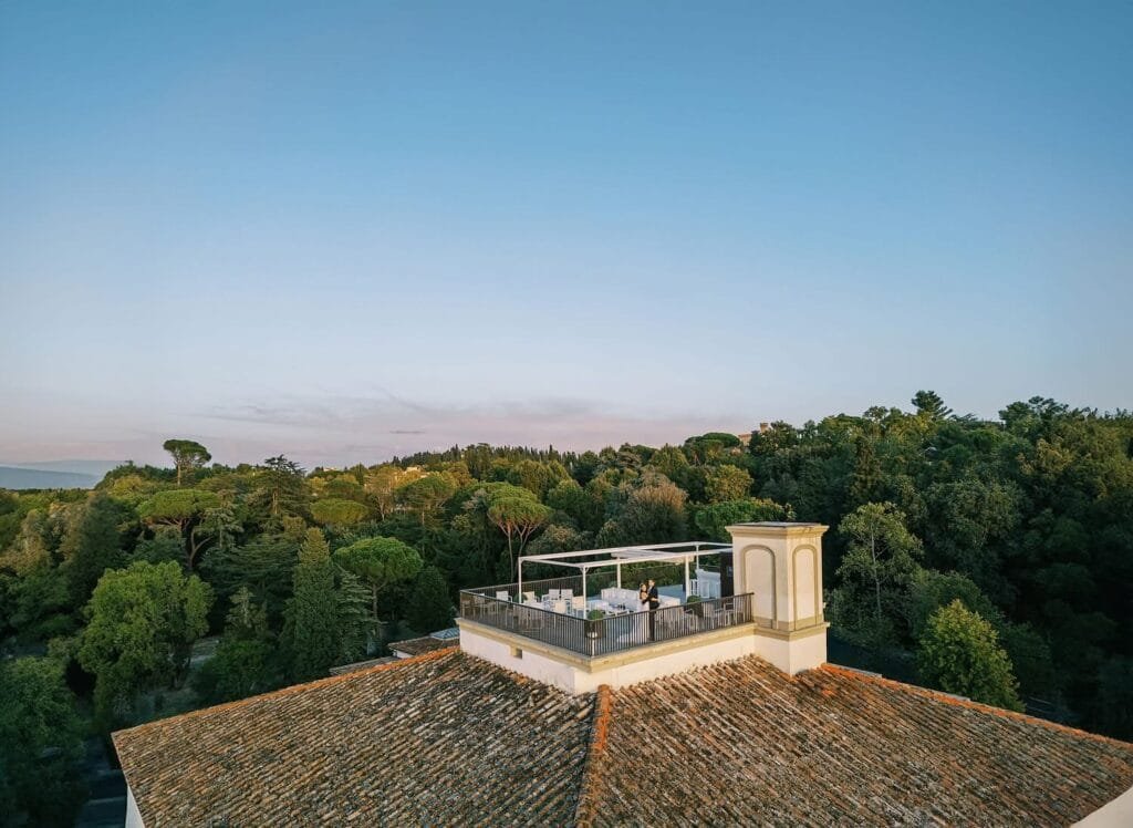 On the rooftop terrace of Villa Cora Florence, a person stands amidst tables and chairs, enveloped by lush green trees under a clear blue sky. The tiled roof and ornate structure at the corner make it an ideal spot for any destination wedding photographer.