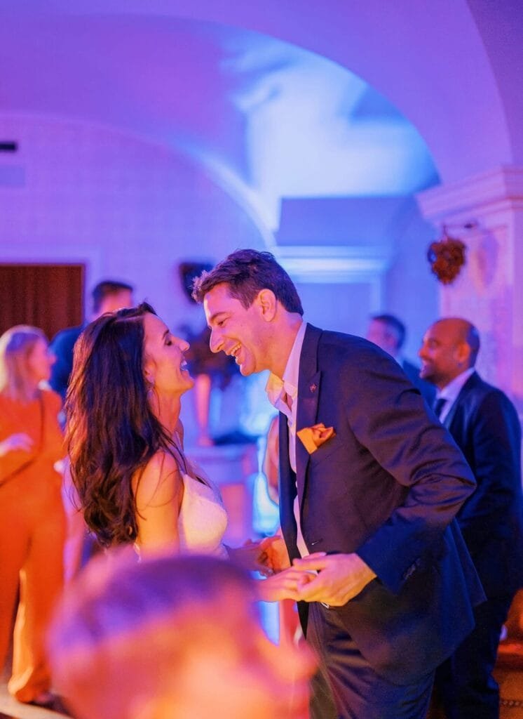 At a Villa Cora destination wedding, a couple joyfully dances amidst elegantly dressed guests. The room glows with colorful lights, enhancing the festive atmosphere. The woman twirls in a white dress while the man complements her in a suit adorned with an orange pocket square.