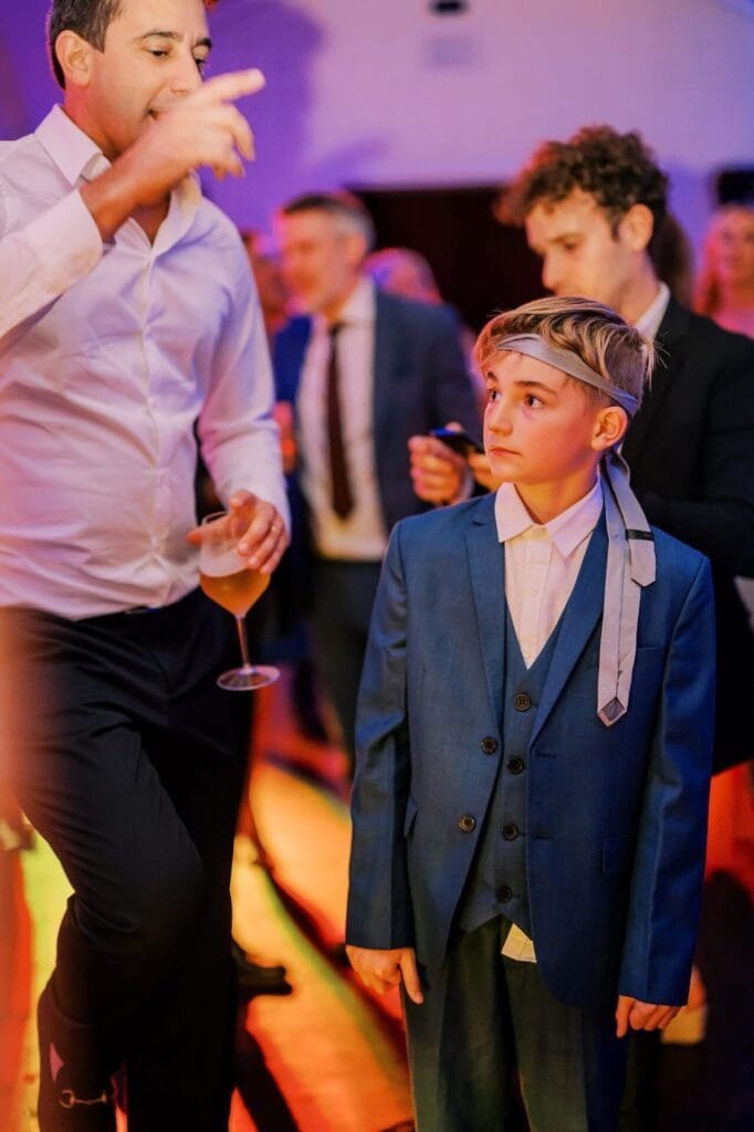 At a vibrant Villa Cora Florence event, a young boy in a blue suit with a tie on his head stands beside a man in a white shirt holding a drink. The boy looks up as the man gestures animatedly. In the colorful lighting, memories are captured by the skilled eye of a destination wedding photographer.