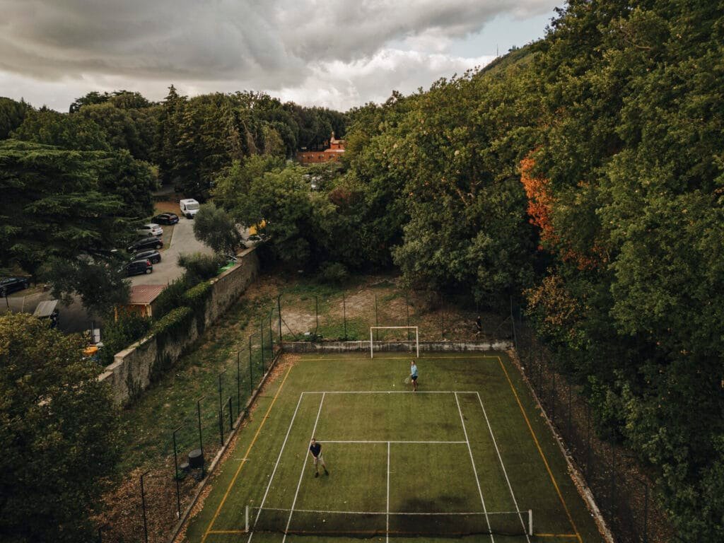 Aerial view of two people playing on a fenced rectangular tennis court at the picturesque Villa Palazzola, nestled among lush green trees and parked cars. The overcast sky with gray clouds adds a dramatic backdrop to this Italian destination near Rome. Perfect for a wedding photographer’s portfolio.