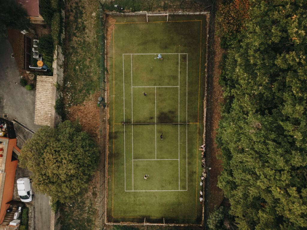 An aerial view captures a rectangular tennis court at Villa Palazzola, an exquisite Italian destination near Rome. Surrounded by lush trees and elegant buildings, several people play energetically while spectators enjoy the match from benches. Nearby, a white car is parked gracefully by the charming surroundings. .