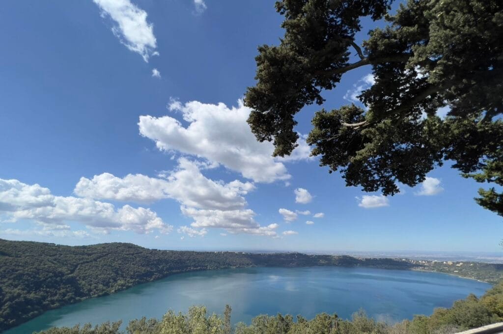 A scenic view of Villa Palazzolas large, blue lake surrounded by lush green hills under a clear blue sky. Fluffy white clouds float above, and tree branches drape over the top right corner, making it an idyllic Italian destination for a Rome wedding photographer.