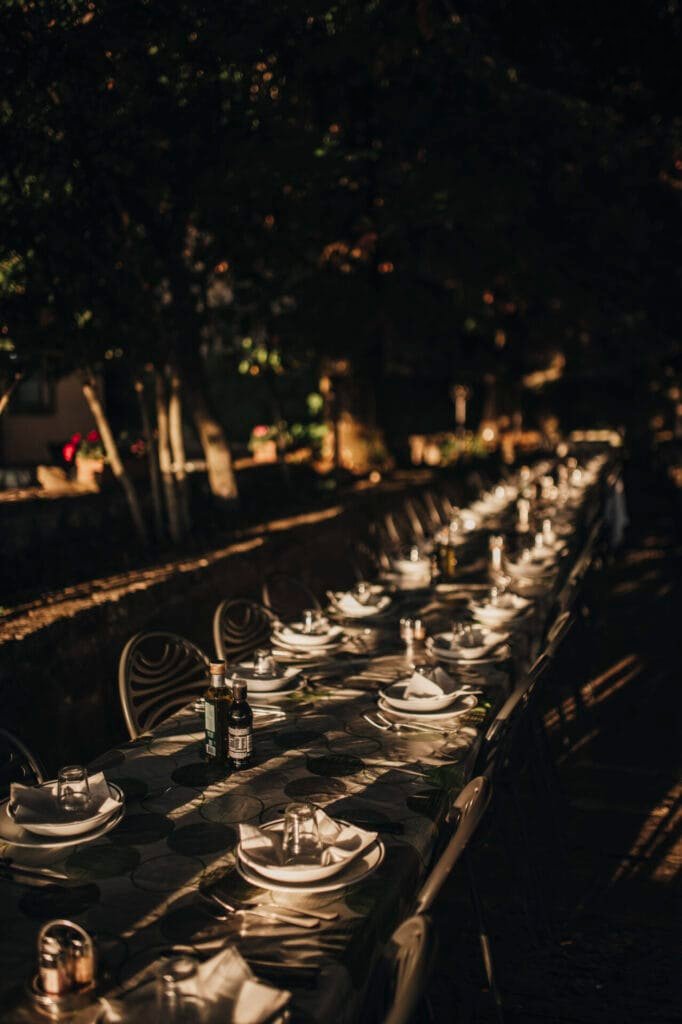 A long outdoor dining table at Villa Palazzola is set for an event, adorned with white plates, folded napkins, candlesticks, and bottles under the dappled sunlight. Trees in the background cast shadows, creating a warm, inviting atmosphere perfect for a destination Rome wedding.