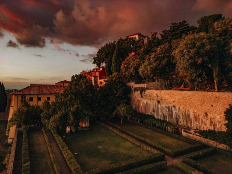 A serene landscape at sunset showcases Villa Palazzola, a historic destination near Rome, with terracotta roofs surrounded by lush trees and manicured hedges. The dramatic clouds cast a warm glow, perfect for any wedding photographer in Italy seeking an idyllic scene.