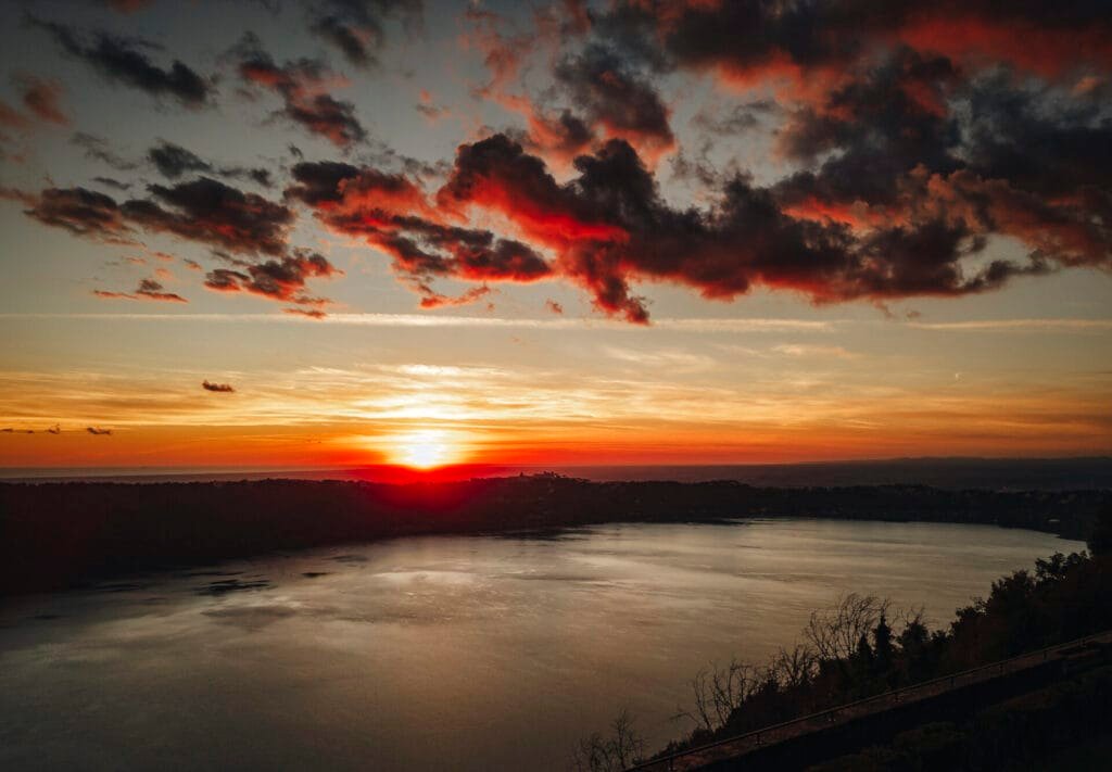 A vibrant sunset over a calm body of water near Villa Palazzola, with the sun partially hidden by the horizon. The sky is painted in shades of orange, red, and blue, complemented by scattered dark clouds. Silhouettes of trees line the edge, creating a picturesque setting for any wedding photographer in Italy.