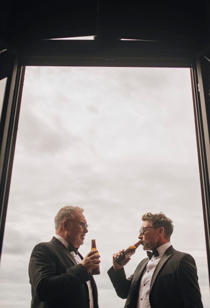 Two men in tuxedos stand near a large open window at Villa Palazzola, each holding a beer bottle. They are smiling and engaged in conversation, with the cloudy sky over Rome visible in the background—a perfect moment captured by a wedding photographer in Italy.