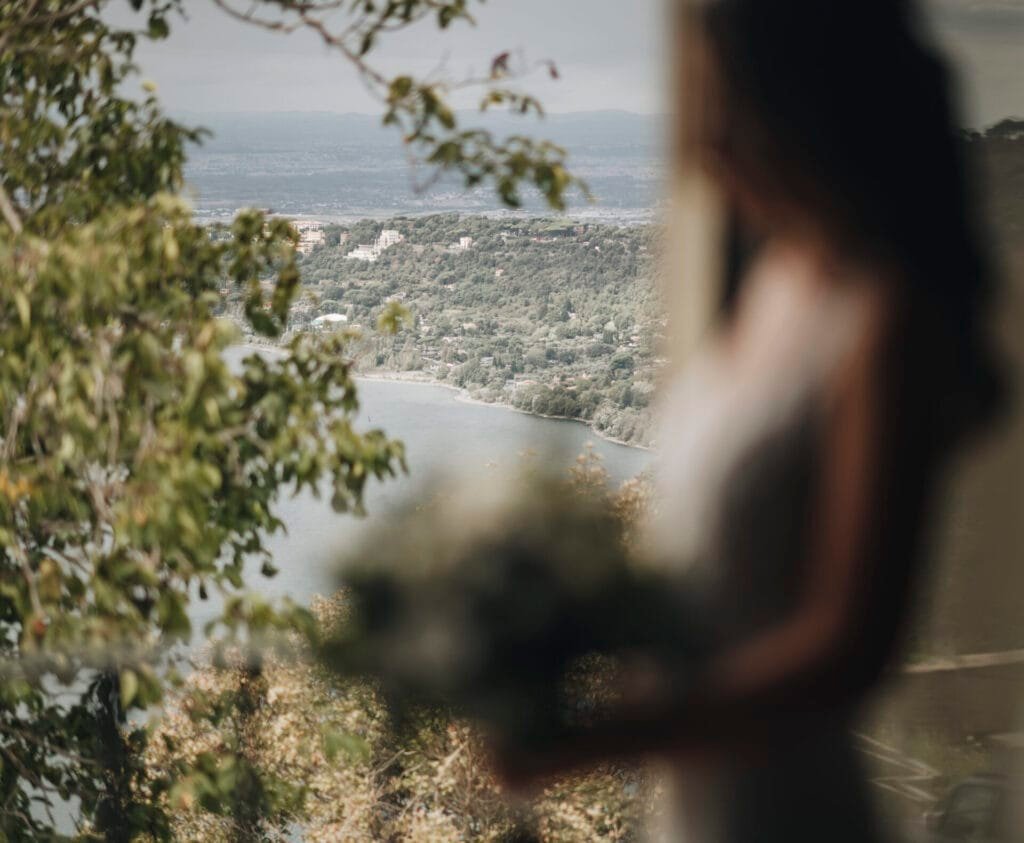 A blurred person stands in the foreground holding flowers, set against a scenic view of Villa Palazzola with its picturesque lake and lush green hills. Trees frame this serene Italian destination near Rome, making it an ideal backdrop for any wedding photographer.