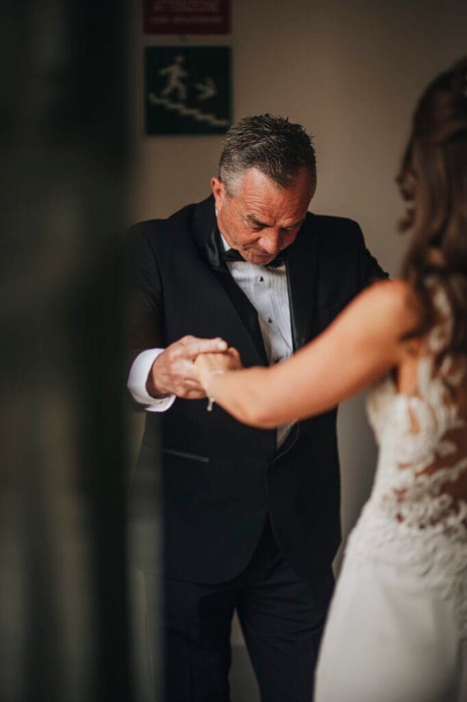 A man in a black tuxedo holds hands with a woman in a white bridal gown at Villa Palazzola in Rome. They share an intimate moment before the ceremony, captured beautifully by their wedding photographer, with the softly blurred background emphasizing their connection.