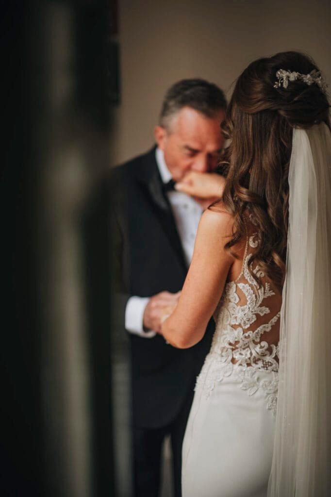 A bride in a lace wedding dress and veil holds hands with an older man in a suit at Villa Palazzola. The man kisses her hand tenderly, capturing a sentimental moment beautifully framed by a talented wedding photographer in Italy, with the background softly blurred.