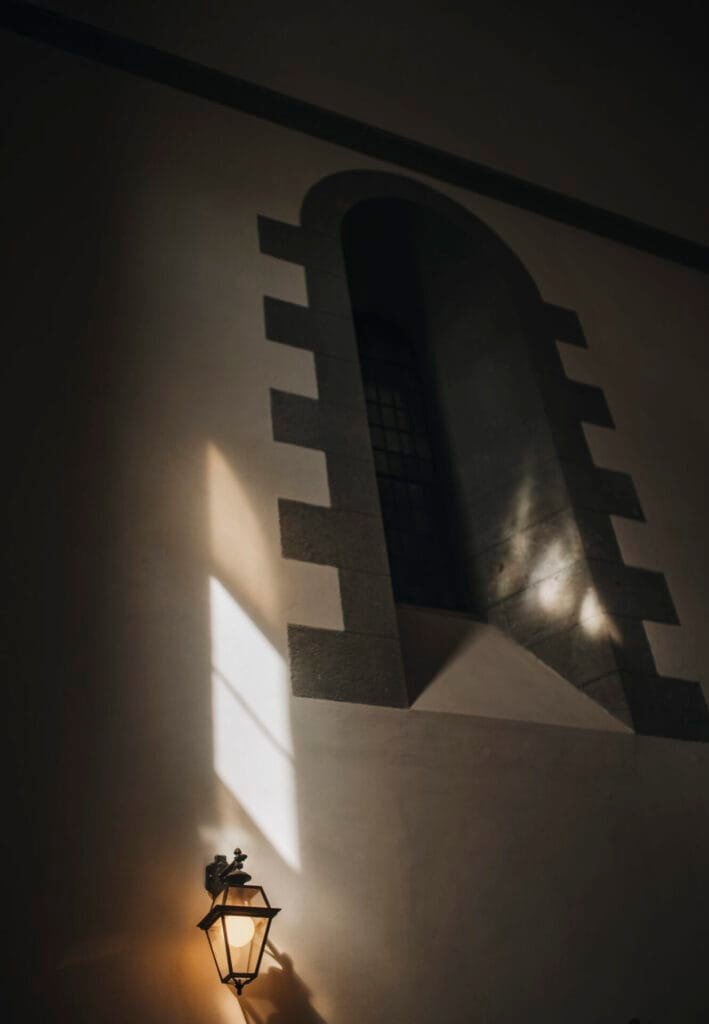 A dimly lit arched window, typical of a Villa Palazzola setting, is framed by stone details on a dark wall. Light streams through the window, creating contrasting shadows and illuminating a vintage wall-mounted lantern below—a perfect scene for an Italian wedding photographer.