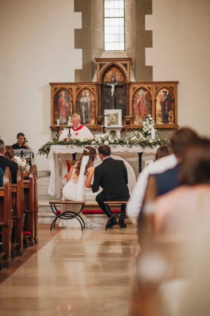 A bride and groom kneel at the altar during a picturesque church wedding ceremony. The priest stands in the background, his white robe adorned with red accents. Surrounded by greenery and flowers, guests fill the pews. Capture these enchanting moments with a Rome wedding photographer in Italy.