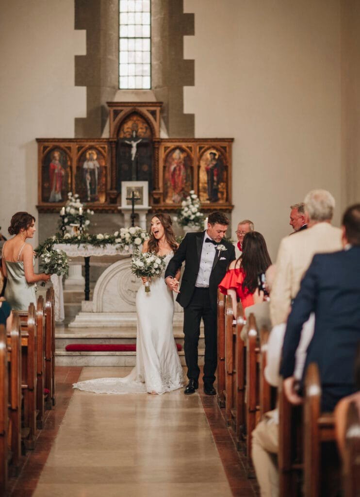 A bride and groom walk joyfully down the aisle of a charming church near Villa Palazzola after their destination wedding. The bride in her white dress and the groom in a black suit are captured beautifully by a talented photographer in Italy, surrounded by smiling guests and elegant floral arrangements.