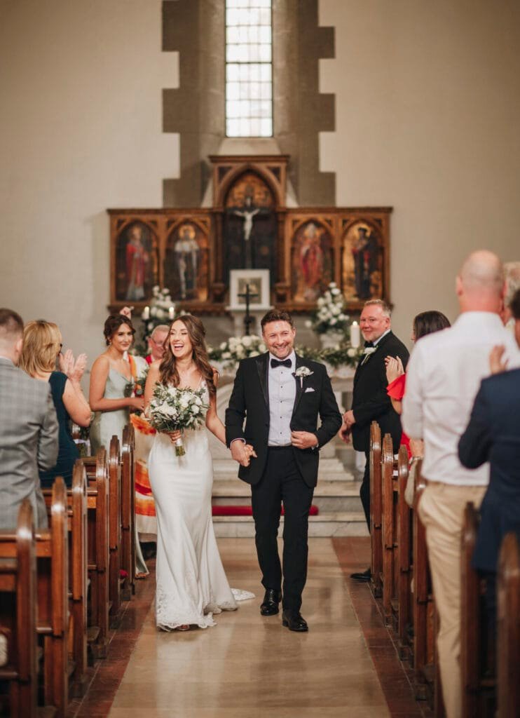 A bride and groom walk hand in hand down the aisle of a charming church near Villa Palazzola, smiling joyfully. The bride is in a white dress with a bouquet, while the groom shines in a black suit with a bow tie. Guests, captured by their wedding photographer in Italy, applaud warmly on either side.