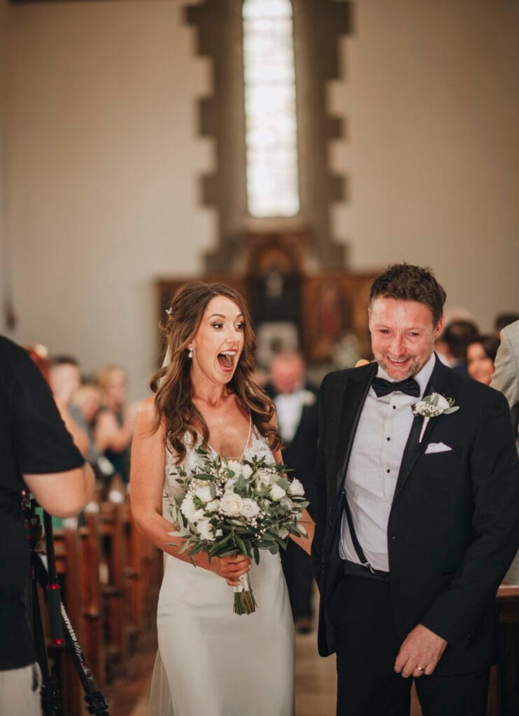 A bride in a white dress holds a bouquet as she walks down the aisle with a man in a black tuxedo, looking surprised and happy. Captured by a talented wedding photographer in Italy, the bright church interior and seated guests complete this unforgettable Villa Palazzola destination wedding near Rome.