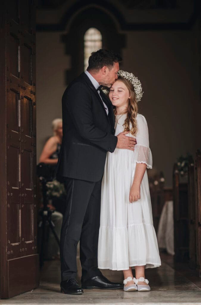 A man in a dark suit lovingly kisses the forehead of a young girl in a white dress and flower crown. They stand in the warmly lit doorway of Villa Palazzola, with pews and a blurred figure in the background. This Rome destination wedding photo captures pure affection and tenderness.