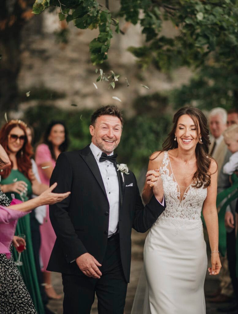 A happy couple walks hand in hand at an outdoor wedding at Villa Palazzola. The bride wears a white dress with lace detailing, and the groom is in a black suit. Guests around them are smiling, with some throwing confetti. Green leaves hang overhead, captured beautifully by our photographer in Italy.