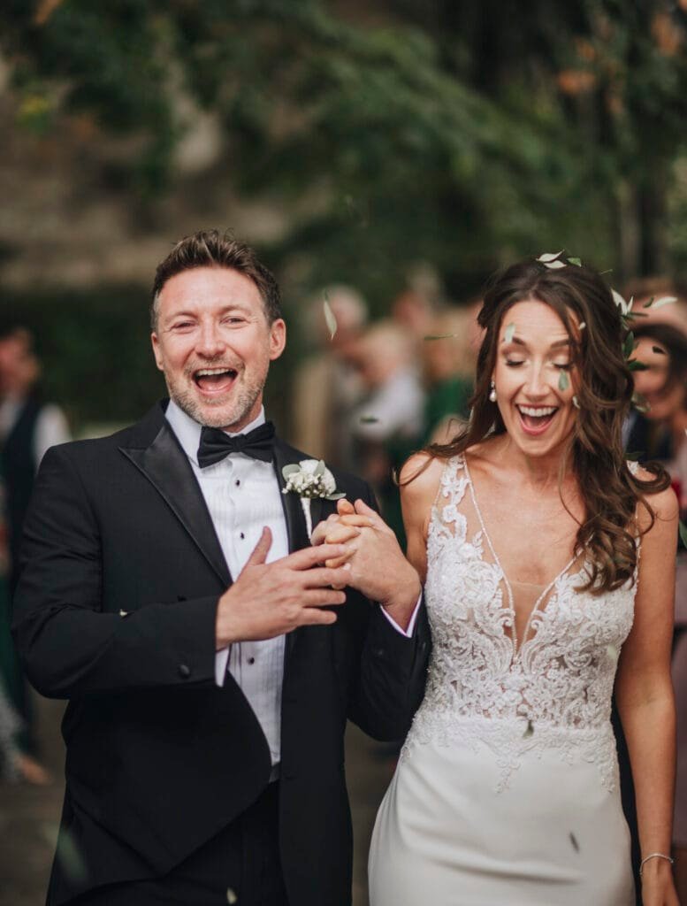 A joyful couple walks hand in hand, wearing wedding attire. The man is in a tuxedo and the woman in a white lace dress. They are smiling, surrounded by blurred greenery at Villa Palazzola. Capturing these moments is their talented Italian Rome wedding photographer.