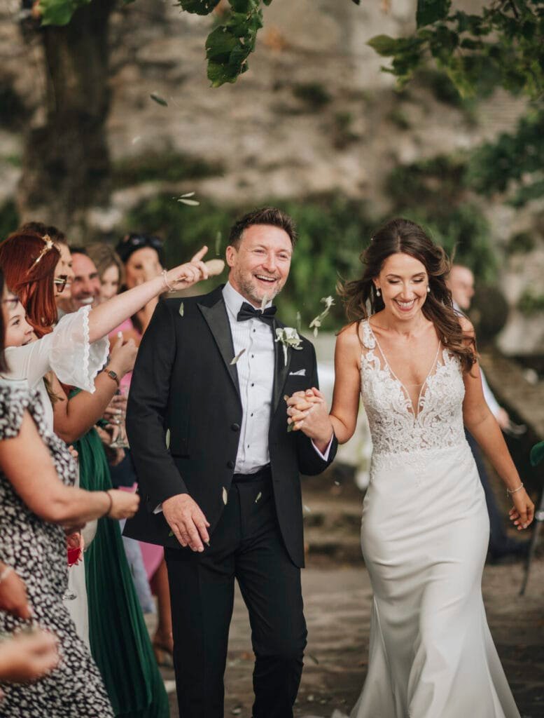 At Villa Palazzola, a bride and groom joyfully walk hand in hand down the outdoor aisle, surrounded by smiling guests. The brides lace wedding dress and the grooms black tuxedo shine under the Italian sun as flower petals fill the air, capturing a perfect moment for any wedding photographer in Rome.