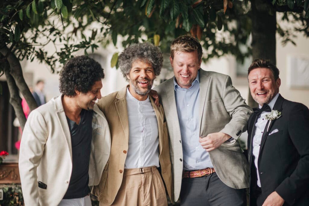 Four men in suits are smiling and standing close together outdoors, one donning a tuxedo. Captured by a Villa Palazzola wedding photographer, they appear to be enjoying the moment, surrounded by greenery in Rome, Italy.