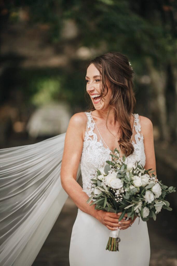 A bride in a white lace wedding dress, holding a bouquet of white and green flowers, smiles joyfully at her Italian Rome wedding. Her hair is styled in loose waves with a flowing veil draped behind her, beautifully captured by the talented Villa Palazzola photographer against a blurred natural setting.