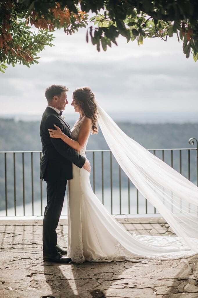 A bride and groom stand facing each other under a tree at Villa Palazzola. The brides long veil flows in the wind as they enjoy the scenic view from the stone terrace. Captured by a talented wedding photographer in Rome, Italy, their special day unfolds under a cloudy sky.