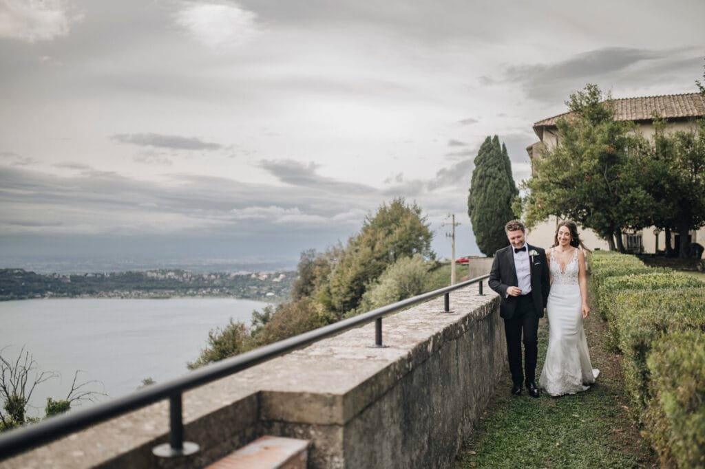 A bride and groom walk along a grassy path next to a stone wall, overlooking a scenic lake near Villa Palazzola. The sky is overcast, with trees and a building visible on the right. Captured by their wedding photographer in Rome, Italy, the couple is dressed in formal wedding attire.