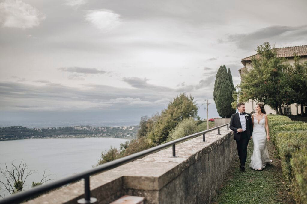 A bride and groom walk along a narrow path lined with greenery, overlooking the idyllic lakeside landscape of Villa Palazzola. The sky is overcast, and the couple, captured by a talented wedding photographer in Rome, Italy, is dressed in elegant formal attire. Tall trees complete the picturesque scene.