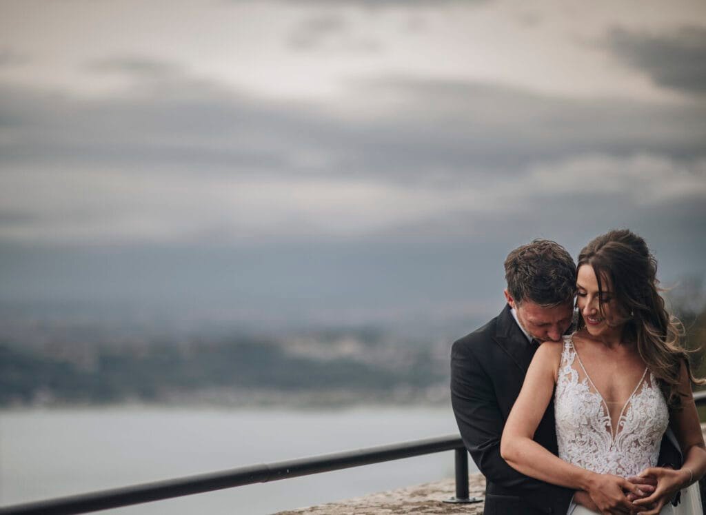 A couple embraces on a balcony at Villa Palazzola, overlooking a scenic view. The woman in a white lace dress smiles as the man in a dark suit hugs her from behind. The background features a cloudy sky and blurred distant landscape, captured beautifully by their wedding photographer in Rome, Italy.