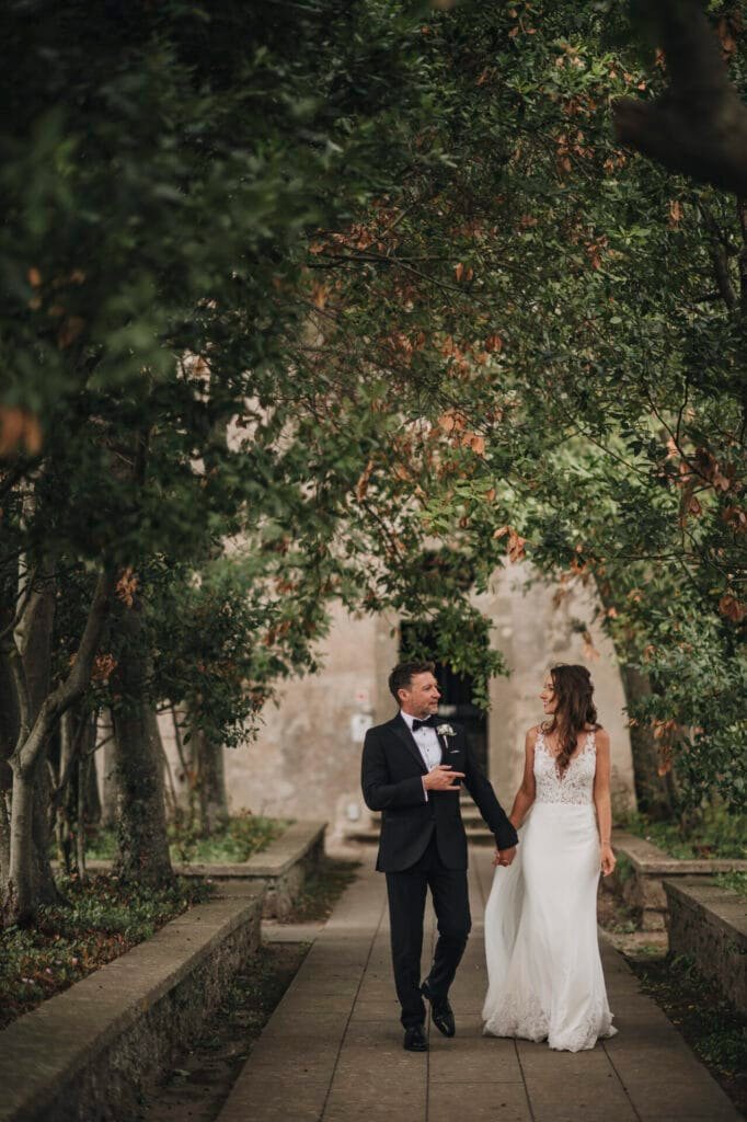 A couple walks hand in hand down a tree-lined pathway at Villa Palazzola, their smiles captured by a wedding photographer in Rome. The woman in her white dress and the man in his black suit are enveloped by lush green foliage, embodying the perfect Italian romance.