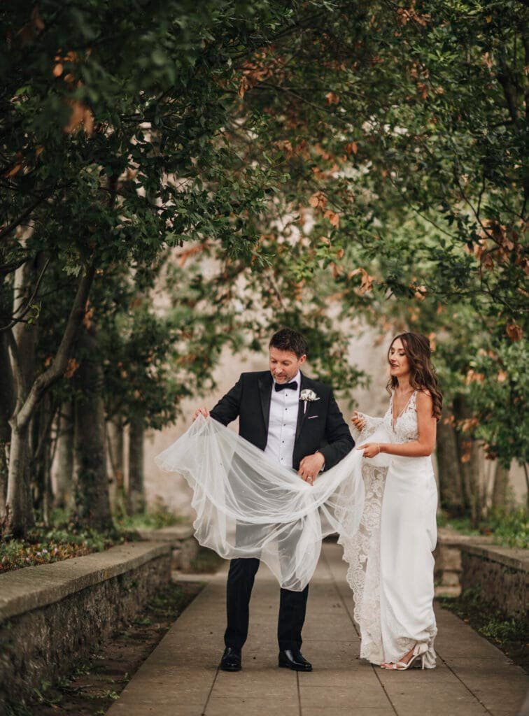 A bride in a white dress and a groom in a black tuxedo stand on a path surrounded by trees at Villa Palazzola. The groom holds part of the brides veil, and she looks at him with a smile. The lush greenery enhances the romantic ambiance, beautifully captured by their wedding photographer in Rome.