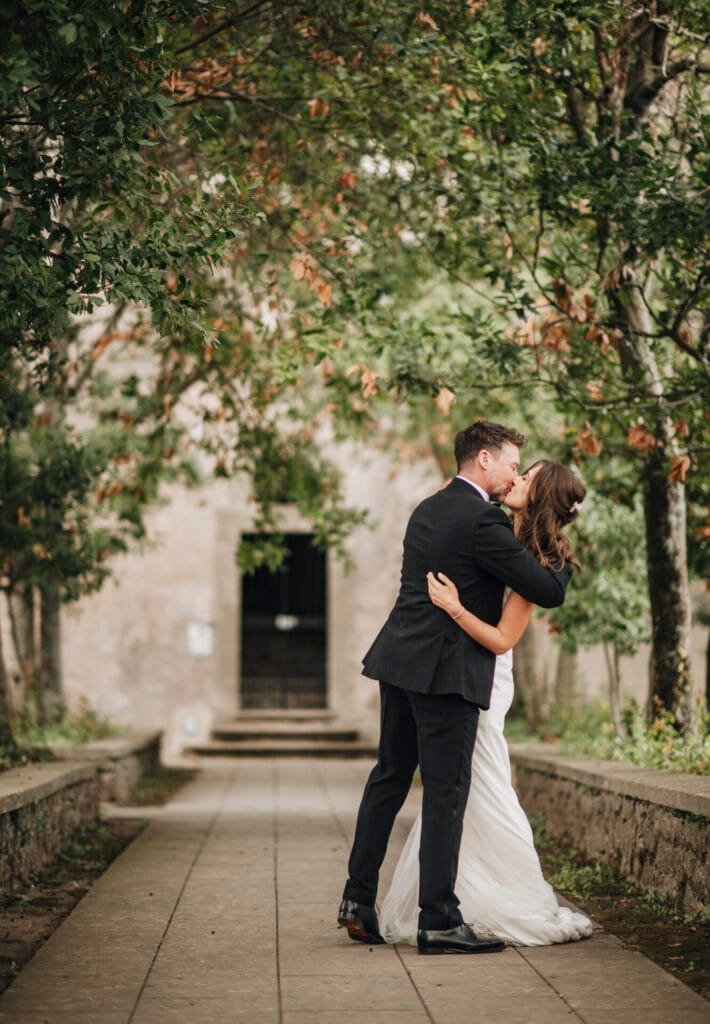 A couple embracing and kissing in a lush garden setting at Villa Palazzola, captured by a wedding photographer in Rome, Italy. The man is in a black suit, and the woman wears a white dress as they stand on a stone path framed by green trees, with an elegant building visible in the background.