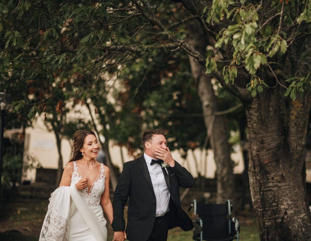 A bride and groom walk hand in hand under tree branches at Villa Palazzola. The bride wears a white dress, the groom in a black tuxedo, both smiling warmly as their wedding photographer captures this beautiful moment in Rome, Italys enchanting outdoor setting.