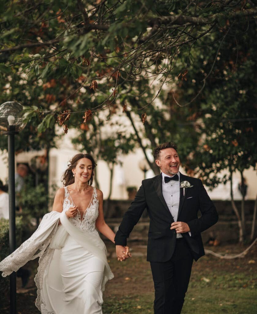 A smiling bride and groom hold hands while walking outdoors, captured beautifully by a Villa Palazzola wedding photographer in Rome. The brides white lace dress and the grooms black tuxedo contrast elegantly against the lush greenery, creating a joyful and celebratory scene.