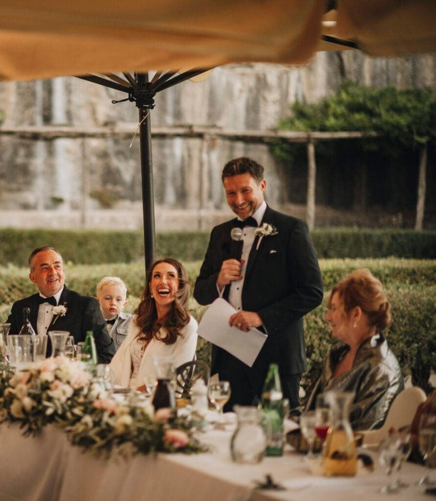 At a Villa Palazzola wedding reception in Rome, Italy, a man in a suit gives a speech with a microphone. A woman in a white dress smiles at him. Others sit nearby, including an older man, a young child, and another woman. The table is elegantly decorated with flowers.