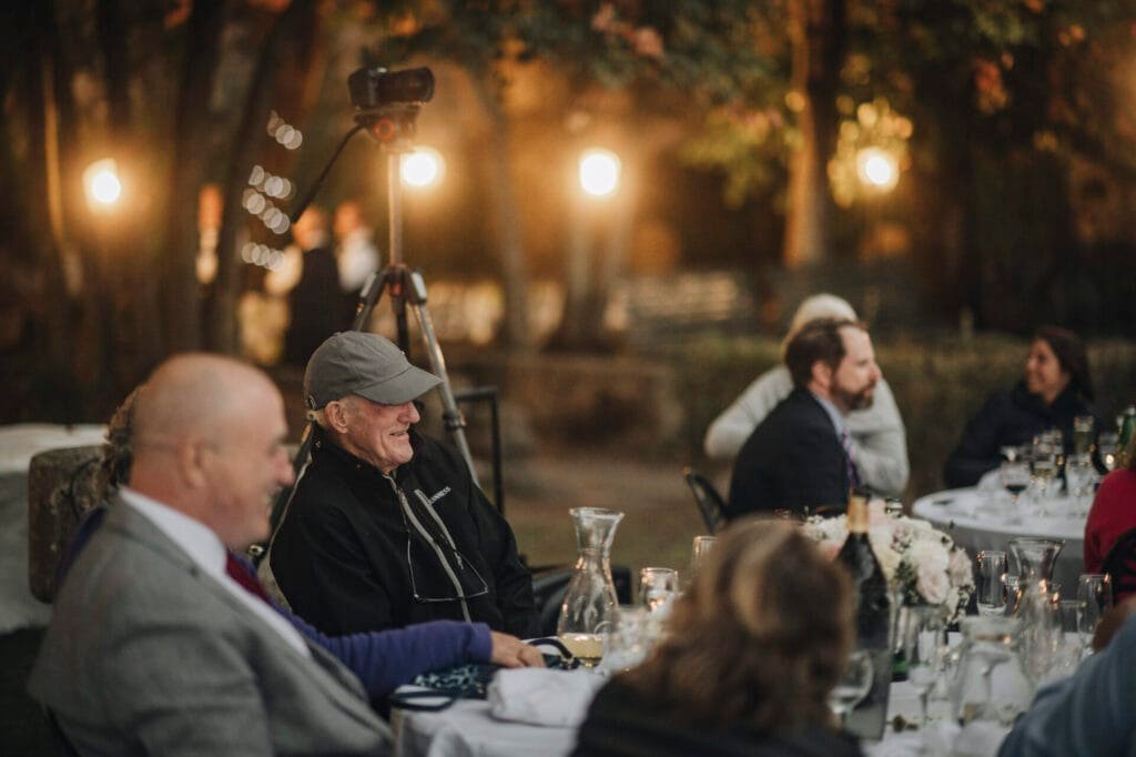 Under the romantic glow of soft lights, guests are seated at round tables adorned with flowers for an evening event. At Villa Palazzola near Rome, a wedding photographer captures moments as a person in a cap stands out amidst diners and chatter, surrounded by lush trees.