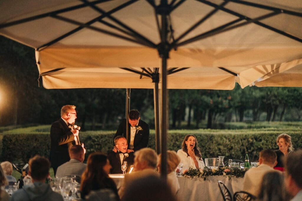 A lively outdoor dinner event at Villa Palazzola in Rome. A person in a suit speaks into a microphone while guests, seated at a long table adorned with greenery and candles, listen and react with smiles and laughter. Trees and soft lighting create a warm ambiance, perfect for capturing by the wedding photographer.