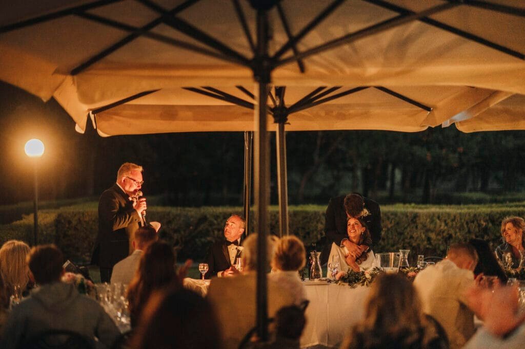 At a charming outdoor wedding reception in Villa Palazzola near Rome, Italy, guests sit under large umbrellas as a person with a microphone addresses the couple at the head table. The warm ambiance, enhanced by soft lighting and lush trees, sets a perfect scene for the wedding photographer to capture.