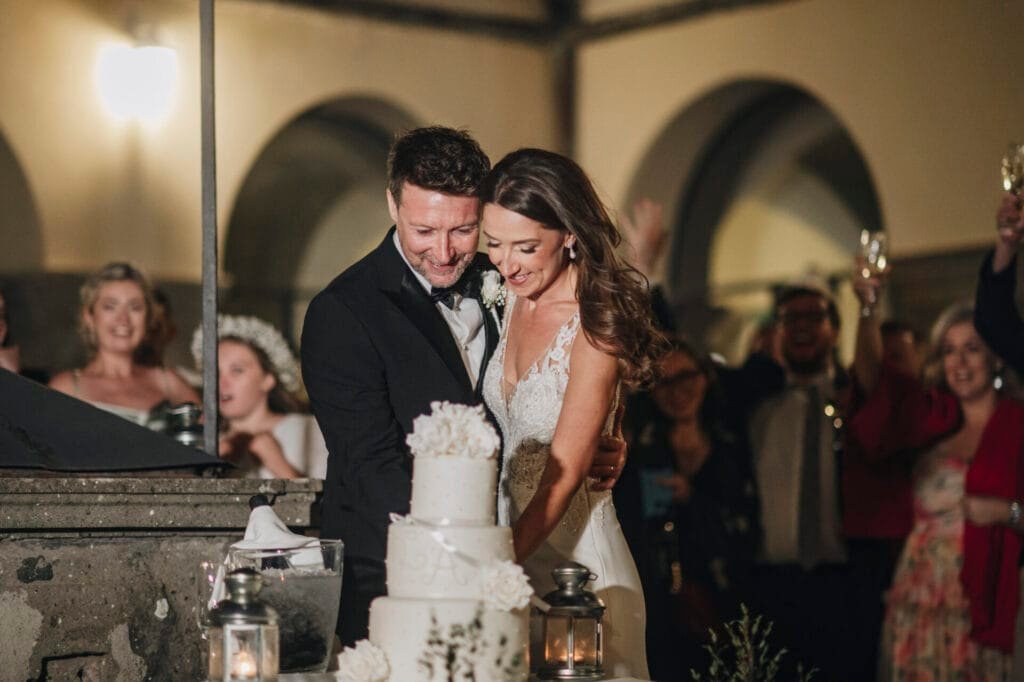 At Villa Palazzola in Rome, a couple in wedding attire joyfully cuts a tiered cake adorned with white floral decorations. Surrounded by cheering guests, some raising glasses in celebration, the warmly lit setting creates a festive and intimate atmosphere perfect for a wedding photographer to capture.