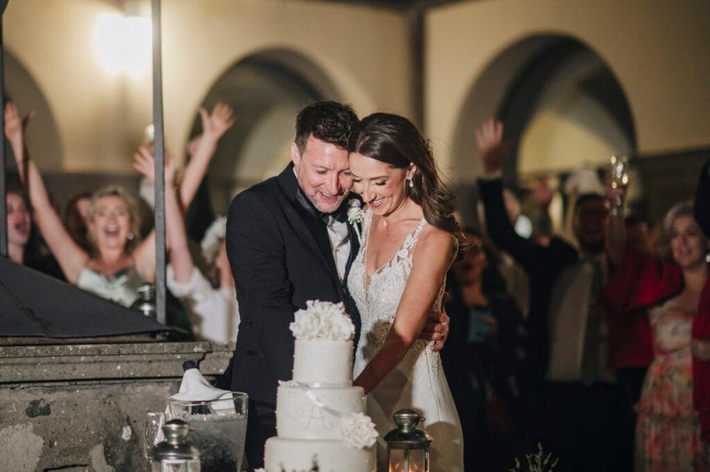 A bride and groom smile as they cut their wedding cake together at a stunning Villa Palazzola venue in Rome, Italy. The multi-tiered cake, adorned with white flowers, stands out as guests cheer and celebrate with arms raised in the background.