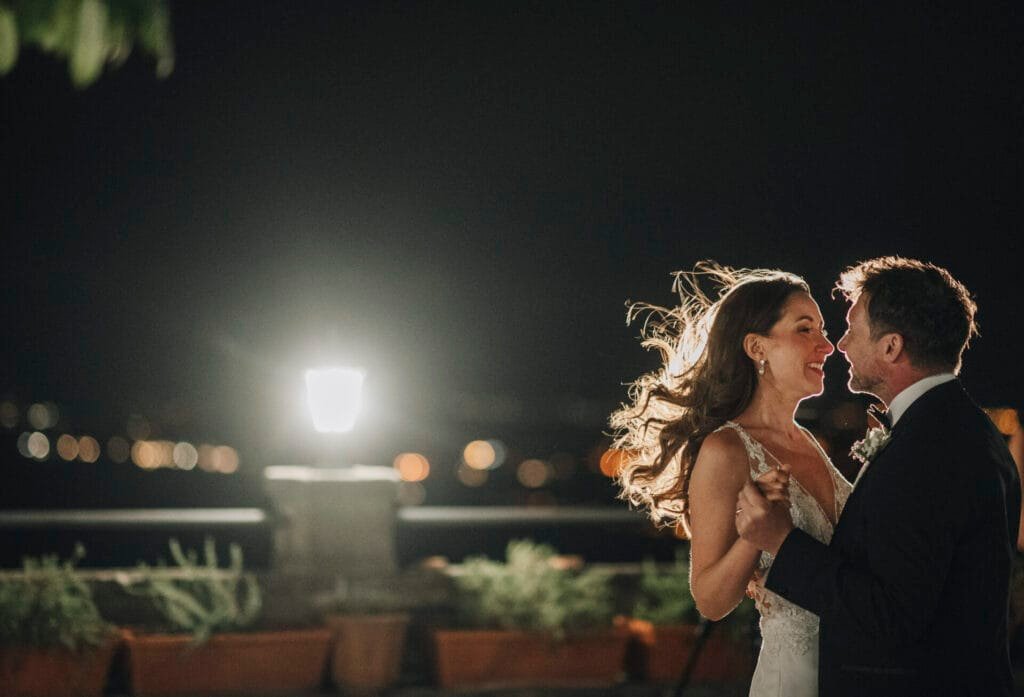 A couple dances joyfully outside at night, illuminated by a soft streetlight. The womans hair flows in the breeze, and both are smiling at each other. The scene captures the romance of a Villa Palazzola wedding photographers dream in Rome, Italy, with a softly lit cityscape in the background.