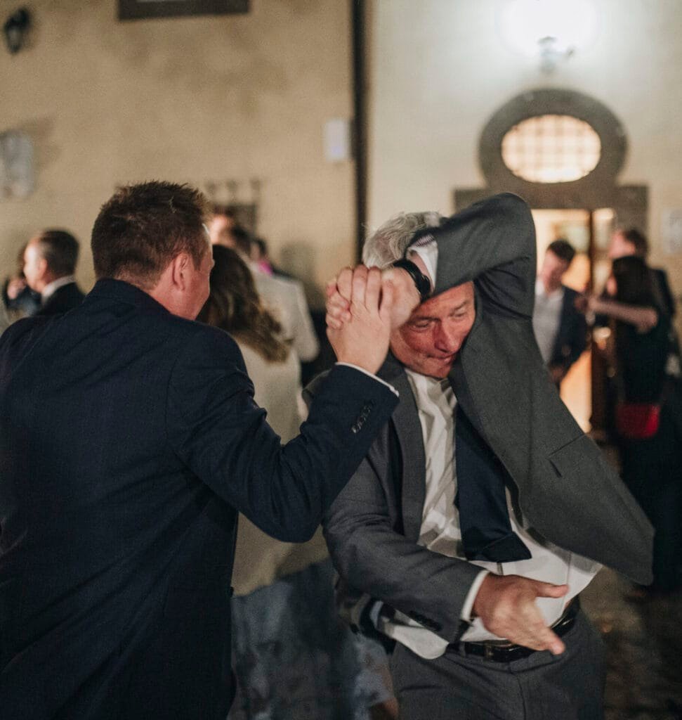 Two men in suits dance energetically at an indoor event at Villa Palazzola. One leads with his arm raised, while the other follows with a playful expression. Blurred people in the background suggest a lively gathering captured beautifully by a wedding photographer in Italy.