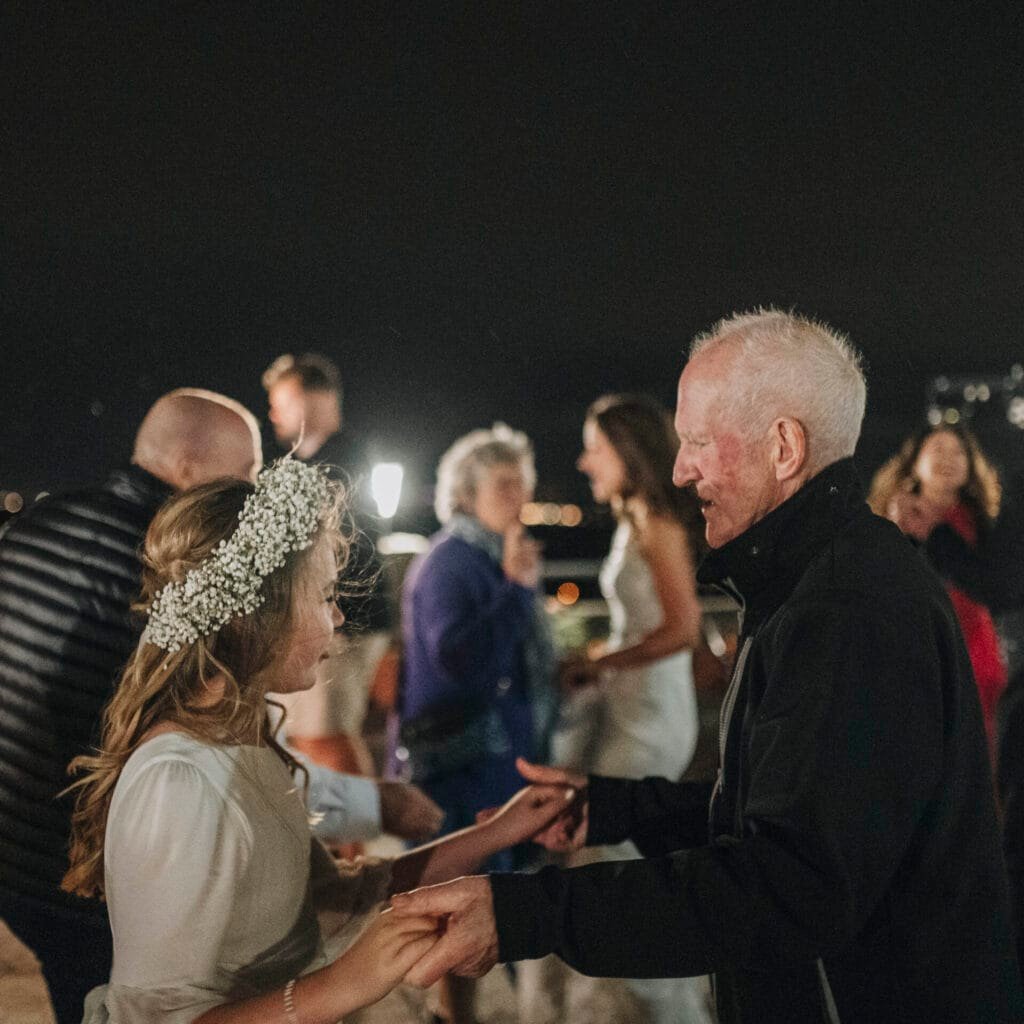 At a picturesque Villa Palazzola wedding in Italy, a young girl in a white dress and floral headband dances gracefully with an elderly man in a black jacket. Amid the blurred background, where several guests twirl under the starlit Roman sky, the joy is beautifully captured by their photographer.