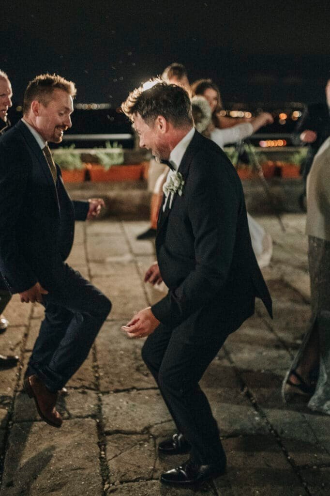 Two men in suits are dancing on an outdoor patio at night. A wedding photographer captures the moment as a woman gleefully takes photos, and another person stands nearby. The festive scene is lively against the backdrop of Villa Palazzolas twinkling lights, perfect for a destination Rome celebration.