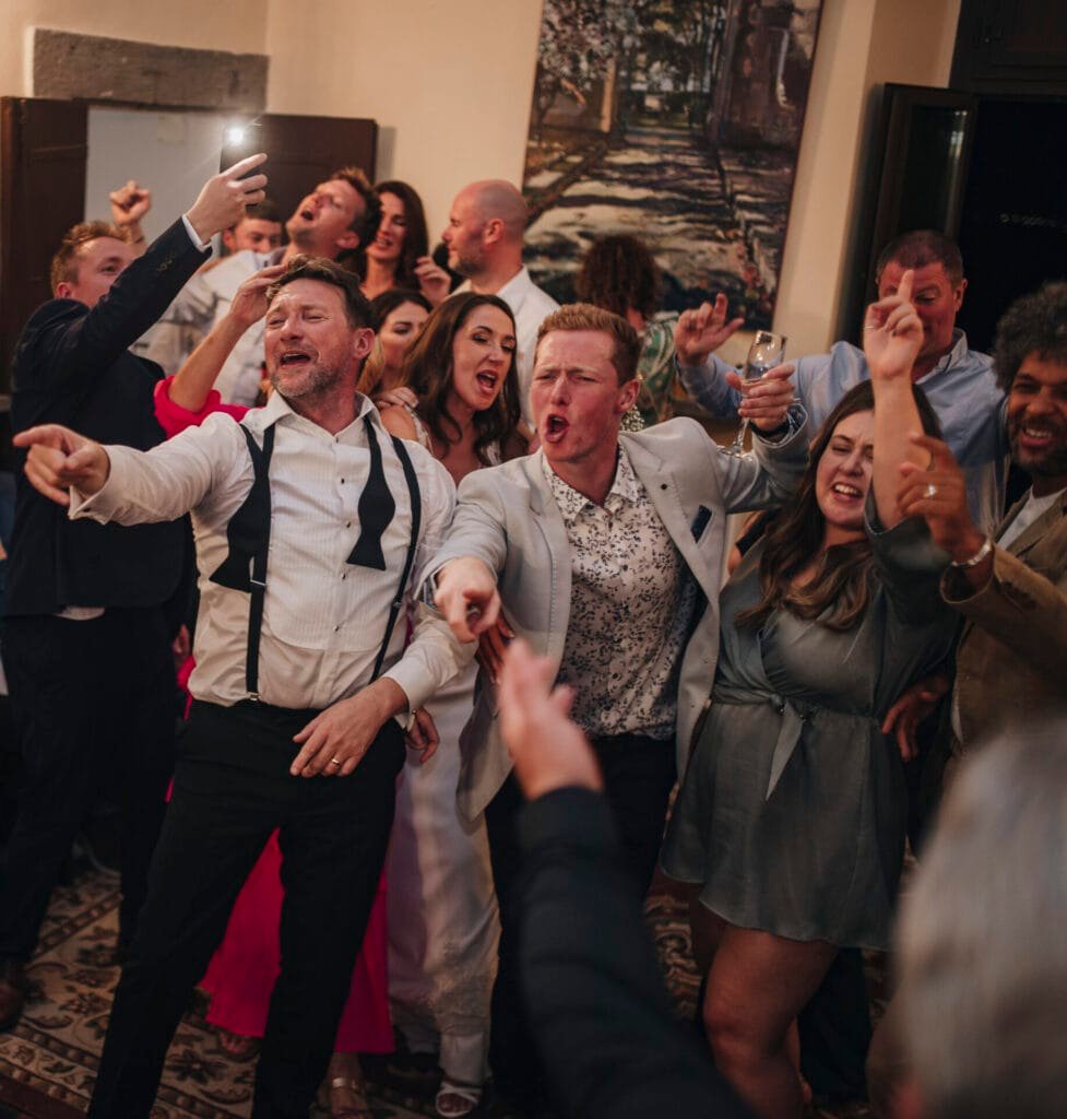 A lively group of people at a Villa Palazzola party in Rome sings and dances. Dressed in formal attire, some hold drinks as the energetic, joyful atmosphere unfolds. One person captures the moment with a selfie, ideal for a wedding photographer in Italy.